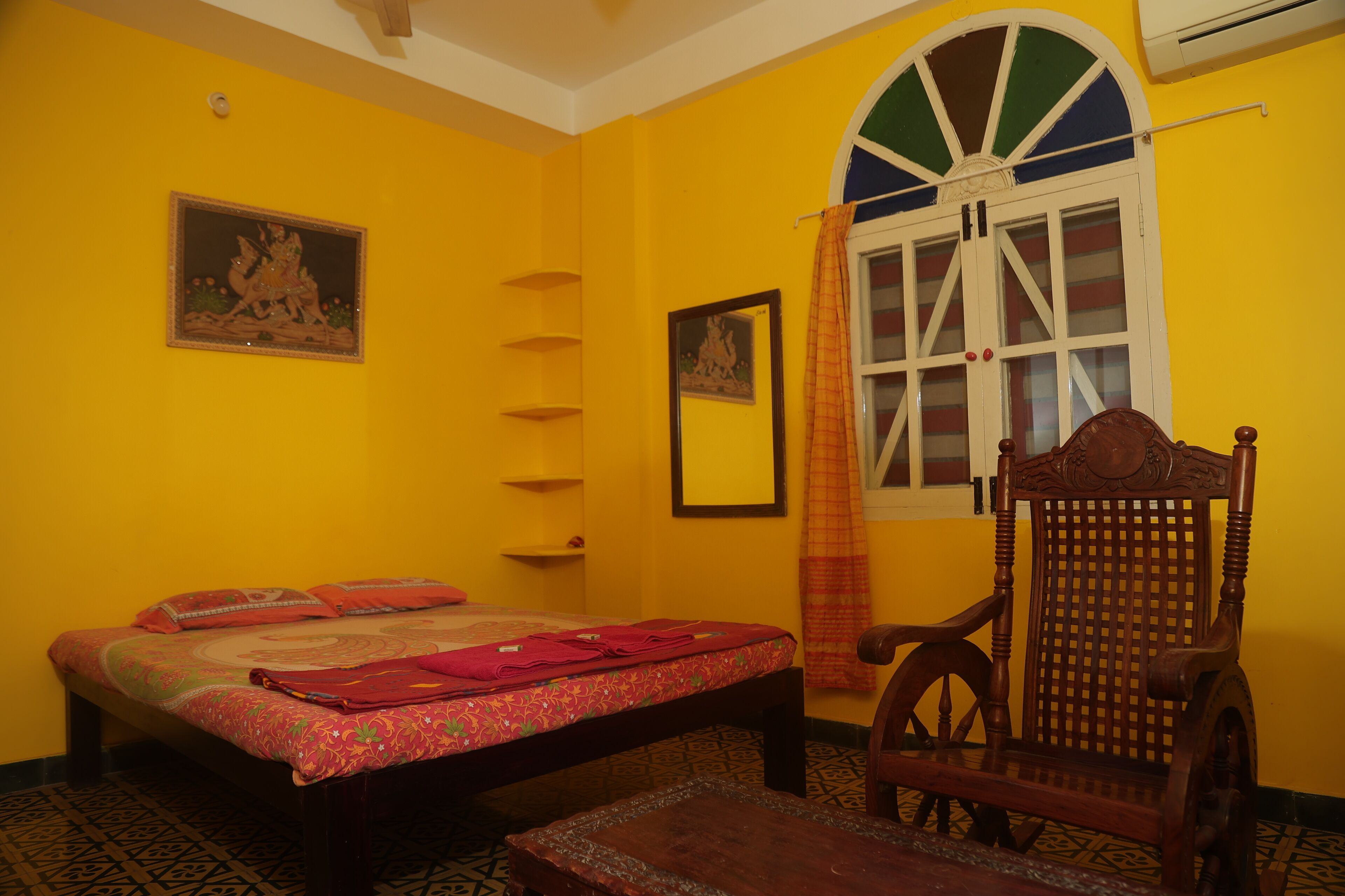A room with a bed, a wooden chair, a window with a curtain, and a wall-mounted shelf.