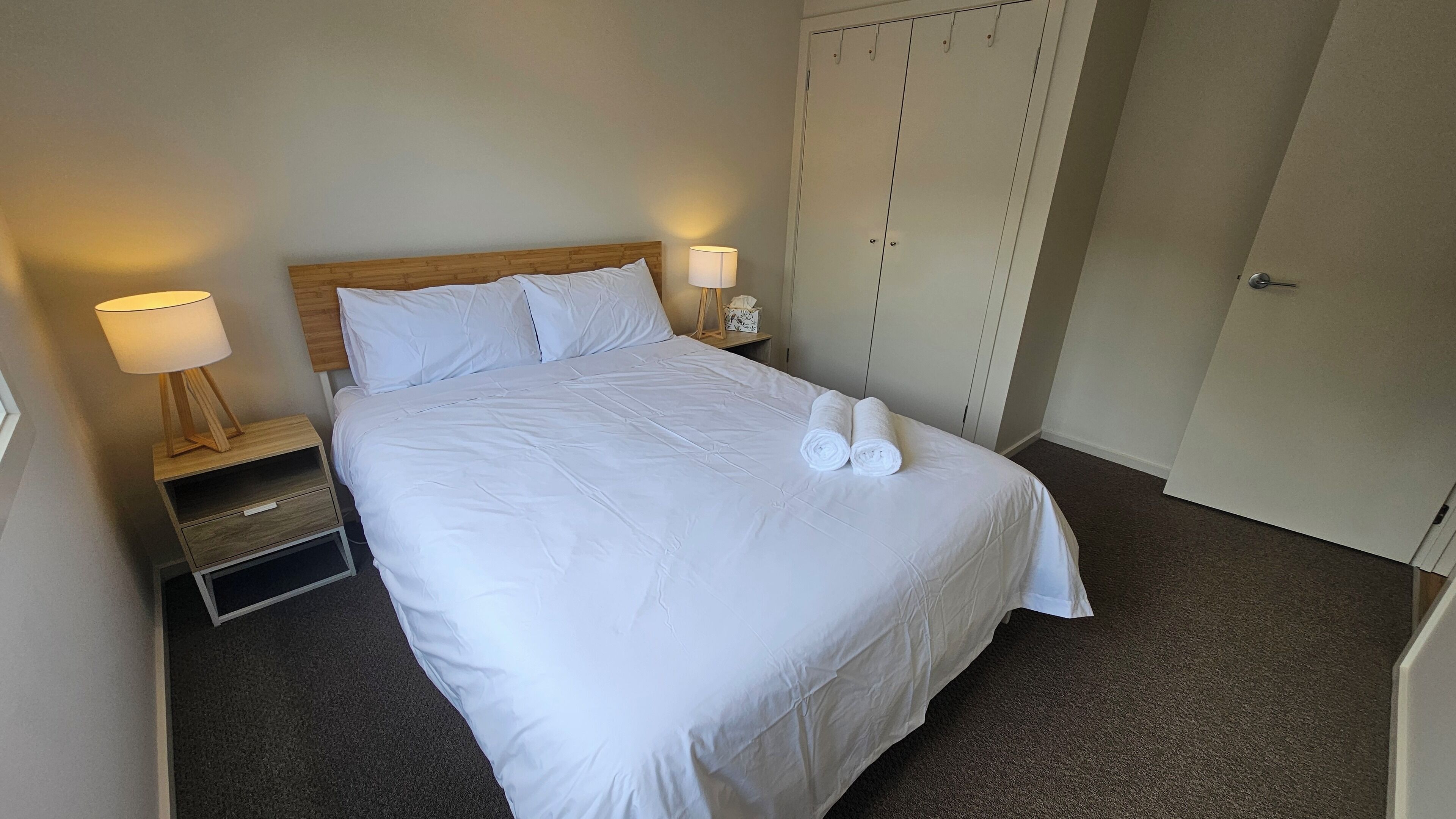 A neatly made bed with white linens, two bedside lamps, a wooden nightstand, and a wardrobe with a mirror.