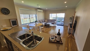 A modern kitchen with a double sink, dining area with a round table and chairs, a sofa, and a view of the outside through large windows. - Getaway on Amaroo Way (Newborough)