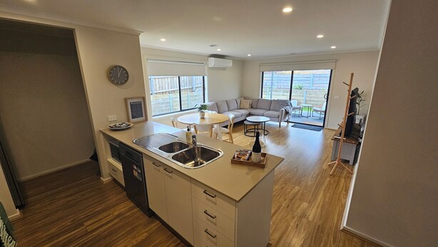 A modern kitchen with a central island, a dining area with a round table, and a living room with a sofa and a TV. - Getaway on Amaroo Way (Newborough)