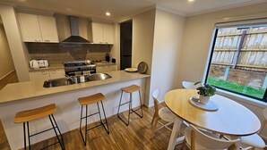 A modern kitchen with a central island, bar stools, a dining table, and a view of a wooden fence through a window. - Getaway on Amaroo Way (Newborough)