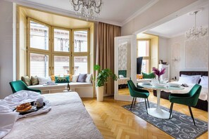 A hotel room with a large window, a dining area with a table and chairs, a bed with a white duvet, and a potted plant.
