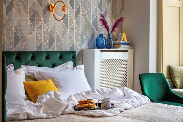 Una cama bien tendida con ropa blanca, un cabecero tapizado en verde y una mesita de noche con objetos decorativos.