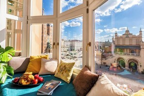 A room with a large window, a view of a city square, and a table with fruits and a book.