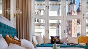 Habitación de hotel con un ventanal grande, una cama con muchas almohadas, una mesita de noche con un vaso y vistas a edificios históricos.