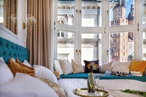 A hotel room with a large window, a bed with multiple pillows, a bedside table with a glass, and a view of historic buildings.