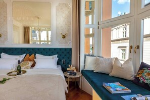 A bedroom with a large bed, a bedside table, a window seat with cushions, and a view of the city.