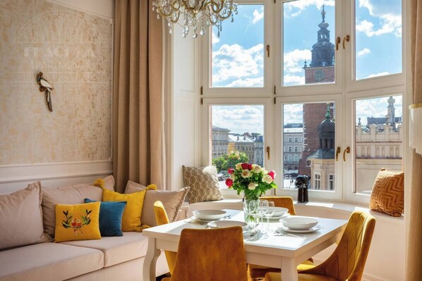 A dining area with a table set for two, a sofa with cushions, a chandelier, and a view of city buildings through large windows.