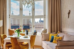 A dining area with a table set for two, a chandelier, and a view of a building through large windows.