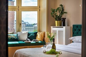 A bedroom with a green sofa, a bed, a window with a view of buildings, a plant on a cabinet, and a tray with grapes.
