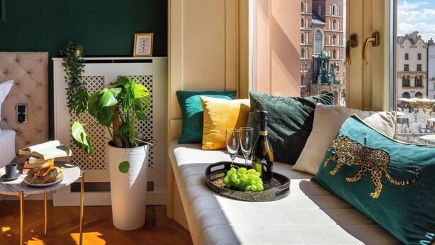 Una habitación elegante con cama, sofá, una mesita con una bandeja de comida y bebidas, una planta en maceta y vistas a un paisaje urbano a través de una ventana.