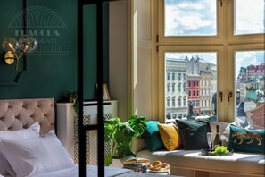 A room with a large window, a tufted headboard, a white cabinet, a plant, a sofa with cushions, and a view of city buildings.