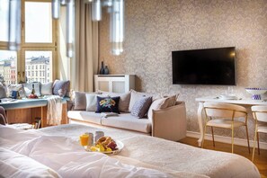 A hotel room with a bed, a sofa, a dining table with chairs, a TV, and a city view.