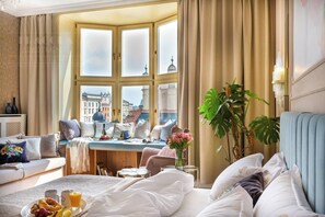 A hotel room with a bay window, a dining area, a bed with white bedding, and a large plant. - Feniks Indigo studio with a Main Square view (Kraków - Feniks)