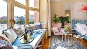 A room with a large window, a sofa with cushions, a chair, a small table with a vase of flowers, and a bed with a blue headboard. - Feniks Indigo studio with a Main Square view (Kraków - Feniks)