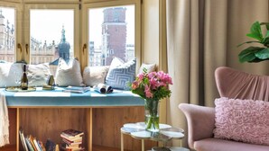 A room with a large window, a seating area with cushions, a small table with a vase of flowers, and a bookshelf. - Feniks Indigo studio with a Main Square view (Kraków - Feniks)