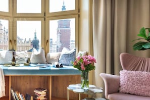 A room with a large window, a seating area with cushions, a small table with a vase of flowers, and a bookshelf. - Feniks Indigo studio with a Main Square view (Kraków - Feniks)