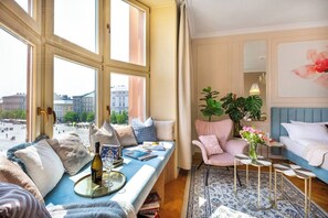 A room with a large window, a sofa with cushions, a chair, a small table with a vase of flowers, and a bed with a blue headboard.