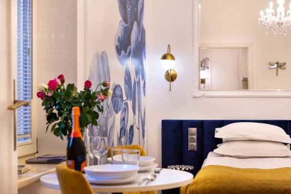 Una habitación de hotel con una cama, una mesa de comedor con platos y una botella, un jarrón de flores y un cuadro floral en la pared.