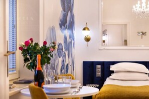 Een hotelkamer met een bed, een eettafel met borden en een fles, een bloevas en een wand met een bloementableau.