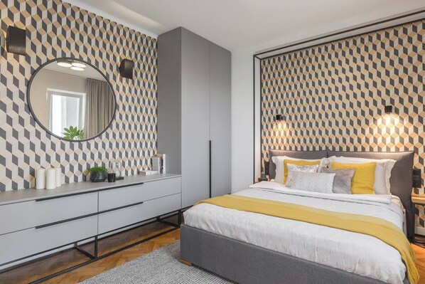 A modern bedroom with a geometric patterned wallpaper, a round mirror, a grey bed with yellow accents, and a bedside table with a lamp. - Feniks Distinguo studio with balcony (Kraków - Feniks)