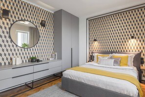 A modern bedroom with a geometric patterned wallpaper, a round mirror, a grey bed with yellow accents, and a bedside table with a lamp.