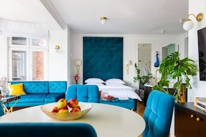 A hotel room with a bed, a blue sofa, a dining table with fruit, and a large window.