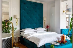 A hotel room with a large teal headboard, a bed with white linens, a small bedside table with a lamp, and a window with a view. - Feniks Jacuzzi Time studio (Kraków - Feniks)
