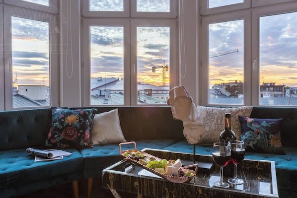 A room with a large window, a sofa with floral cushions, a bust sculpture, and a tray with snacks and wine glasses.