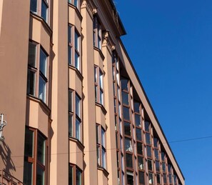 Monikerroksinen rakennus, jonka julkisivu on beige, siinä on suuret ikkunat ja taivaalla on kirkas sininen taivas.