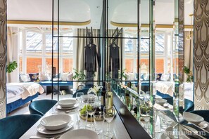 A dining area with a long table set for a meal, a view of a bedroom with a bed and a hanging coat rack, and a large window with a view of buildings.