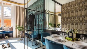 A modern dining area with a glass partition, a blue upholstered chair, a white table set for dining, and a view of the dining room through the partition.