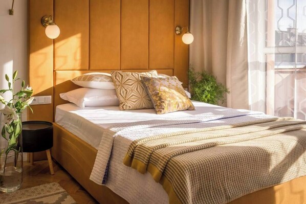 A bed with a wooden headboard, multiple pillows, a bedside table, a vase with flowers, and a window with curtains. - Feniks Sunblush studio with terrace (Kraków - Feniks)