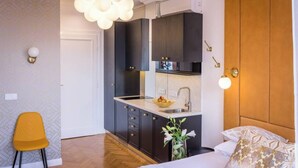 Kompakti hotellihuone, jossa on pieni keittiönurkkaus, kuviollisilla tyynyillä varustettu sänky, keltainen tuoli ja moderni valaisin.
