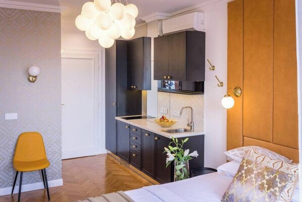 A compact hotel room with a kitchenette, a bed with patterned pillows, a yellow chair, and a modern light fixture. - Feniks Sunblush studio with terrace (Kraków - Feniks)