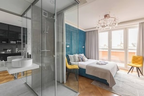 Une chambre d’hôtel moderne dotée d’une douche en verre, d’un grand lit avec un mur d’accent bleu, d’une chaise jaune et d’une vue sur la ville.