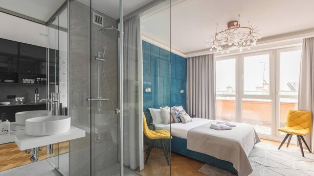 Habitación de hotel moderna con cabina de ducha de vidrio, una cama grande con una pared de acento azul, una silla amarilla y vistas de la ciudad.
