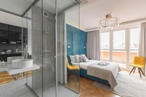Une chambre d’hôtel moderne dotée d’une douche en verre, d’un grand lit avec un mur d’accent bleu, d’une chaise jaune et d’une vue sur la ville.