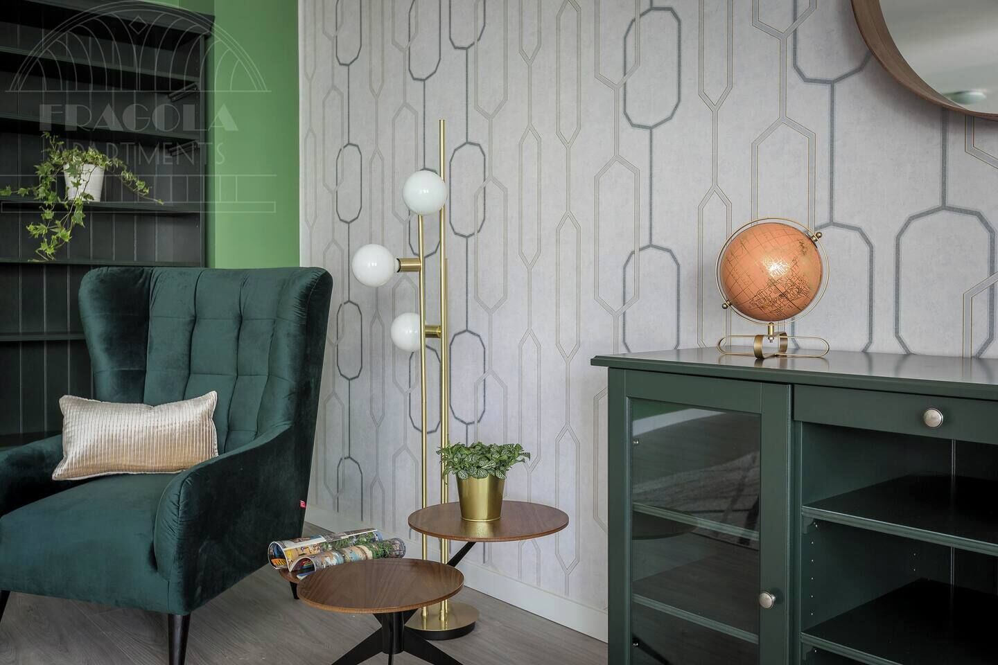 A room with a green armchair, a globe on a desk, a floor lamp, and a patterned wallpaper.