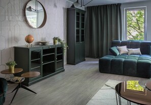 A living room with a dark green sofa, a round mirror, a cabinet with glass doors, and a window with curtains.