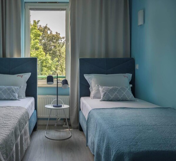 A hotel room with two beds, a small table, a window with a view of trees, and a radiator. - Apartment Serene 3-bed (Kraków – Old Town)