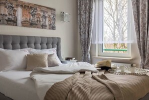 Una cama bien hecha con cabecera tapizada en gris, una mesita de noche con libros y una bandeja con tazas, y una ventana con cortinas.