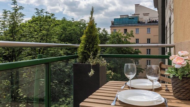 Un balcón con una mesa para dos, una planta en maceta y vistas a edificios y árboles.