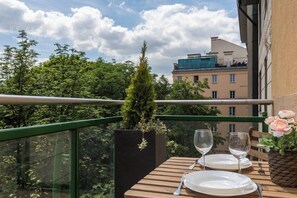 Un balcón con una mesa para dos, una planta en maceta y vistas a edificios y árboles.