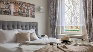 Una cama bien hecha con cabecera tapizada en gris, una mesita de noche con libros y una bandeja con tazas, y una ventana con cortinas.