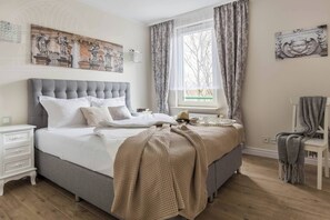Une chambre à coucher avec un grand lit, une table de chevet, une chaise, une fenêtre avec des rideaux et des œuvres d’art murales.