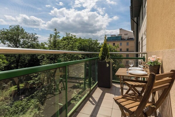 Un balcon avec une table dressée pour le dîner, un pot de fleurs et une vue sur les bâtiments et les arbres environnants.