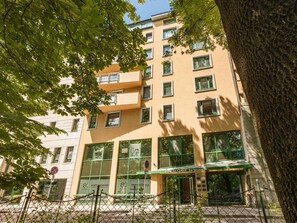 Un edificio de varios pisos con toldo verde, ventanas amplias y un árbol en primer plano.