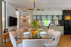 A modern dining room with a glass table, six chairs, a vase of flowers, and a kitchen in the background.