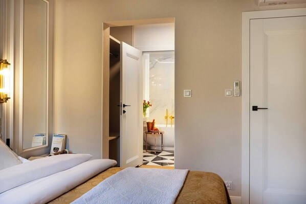 A hotel room with a bed, a mirror, a door, and a view into another room with a tiled floor and a plant.